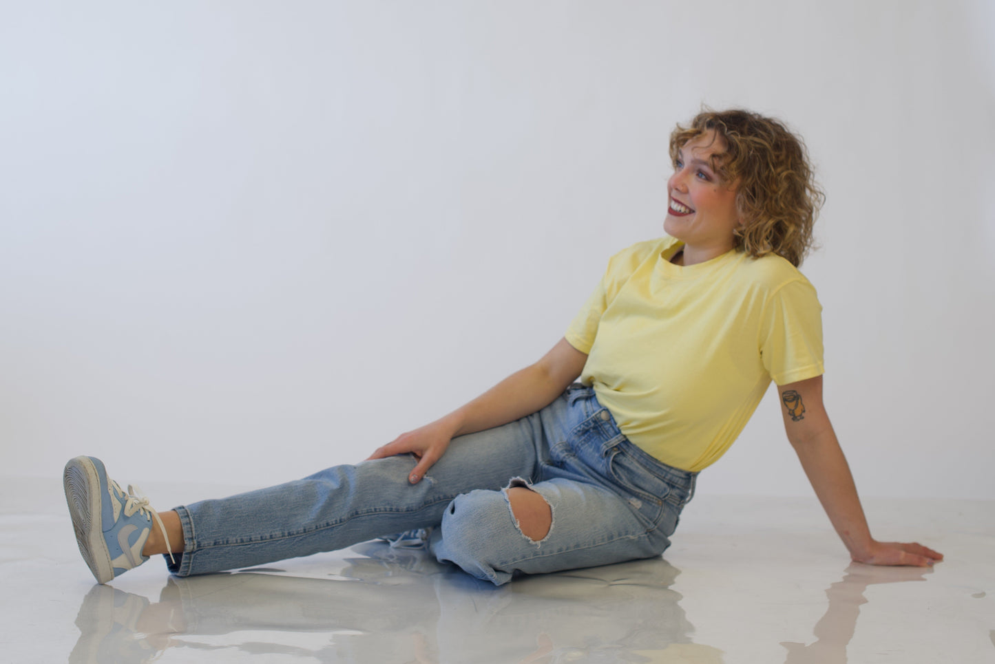 Person sitting on a white floor wearing a yellow shirt and blue jeans.