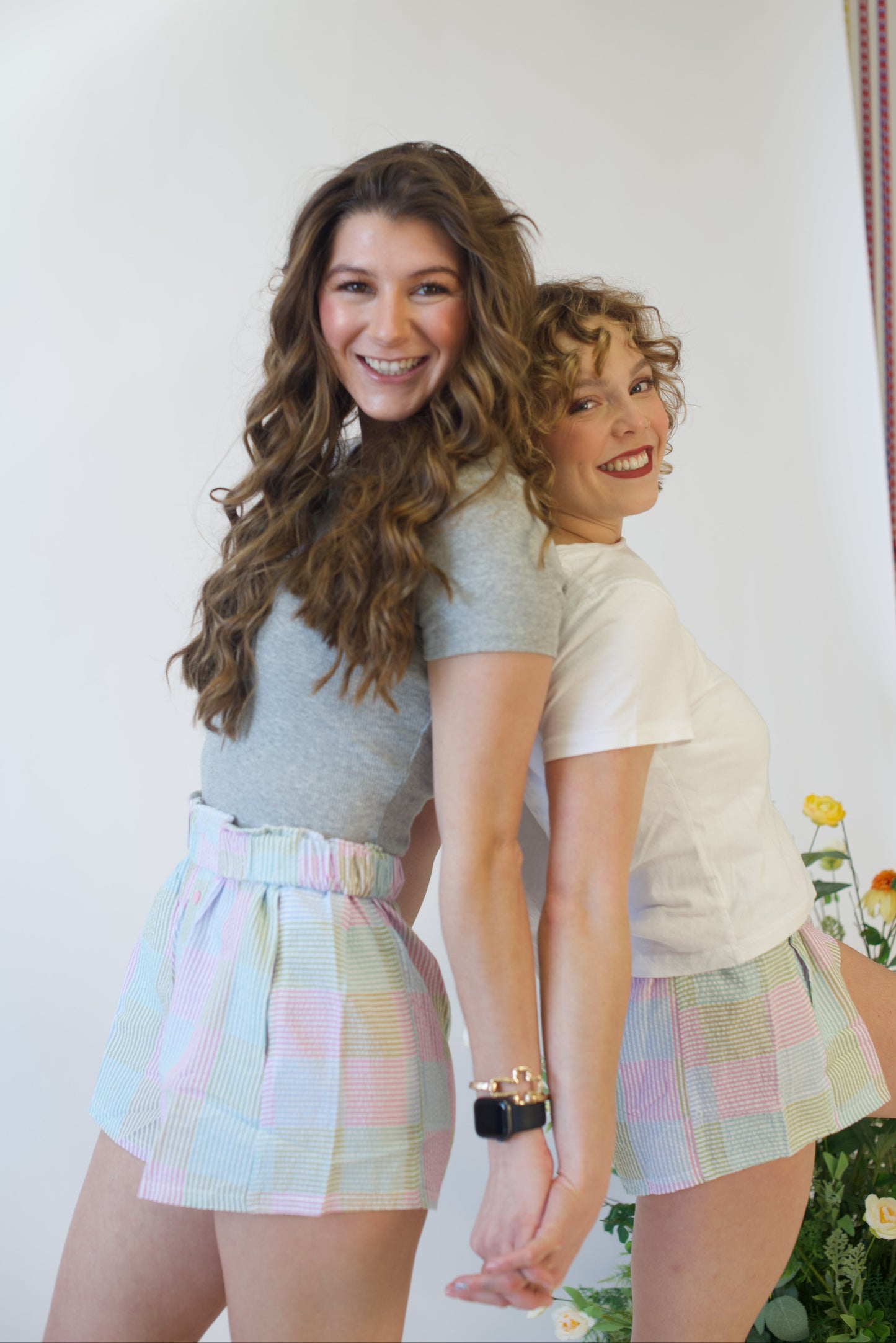 Two women standing close together, smiling, wearing matching pastel checkered shorts and casual tops.