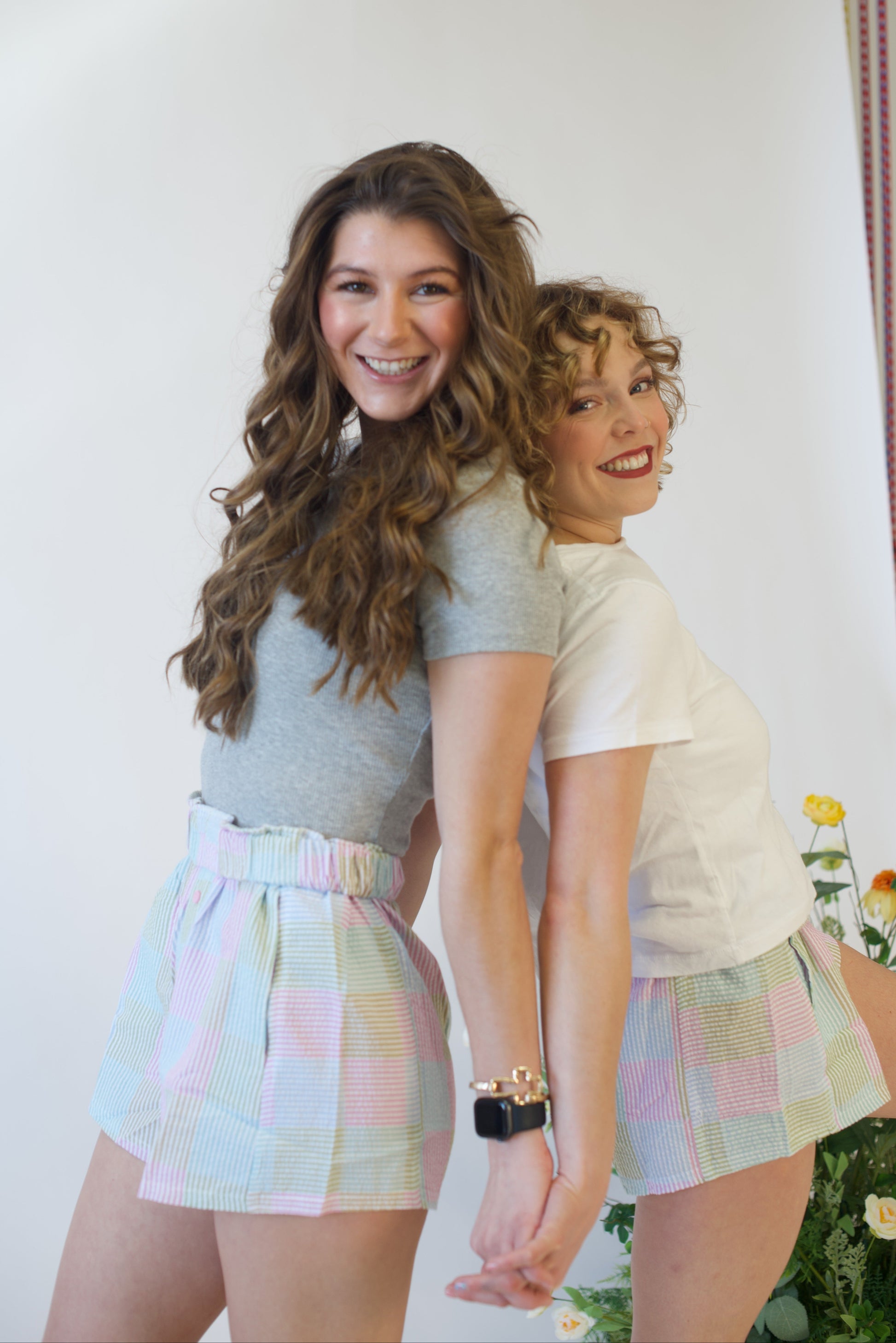 Two women standing close together, smiling, wearing matching pastel checkered shorts and casual tops.