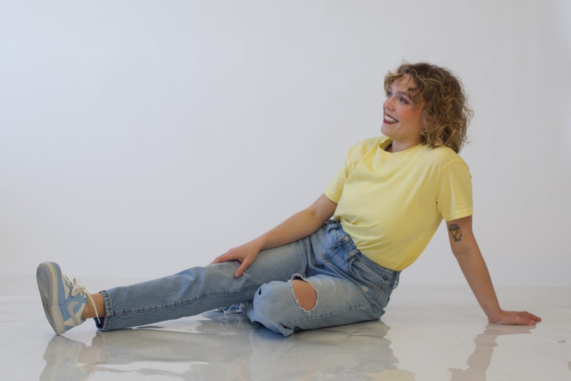 Person sitting on a white floor wearing a yellow shirt and blue jeans.