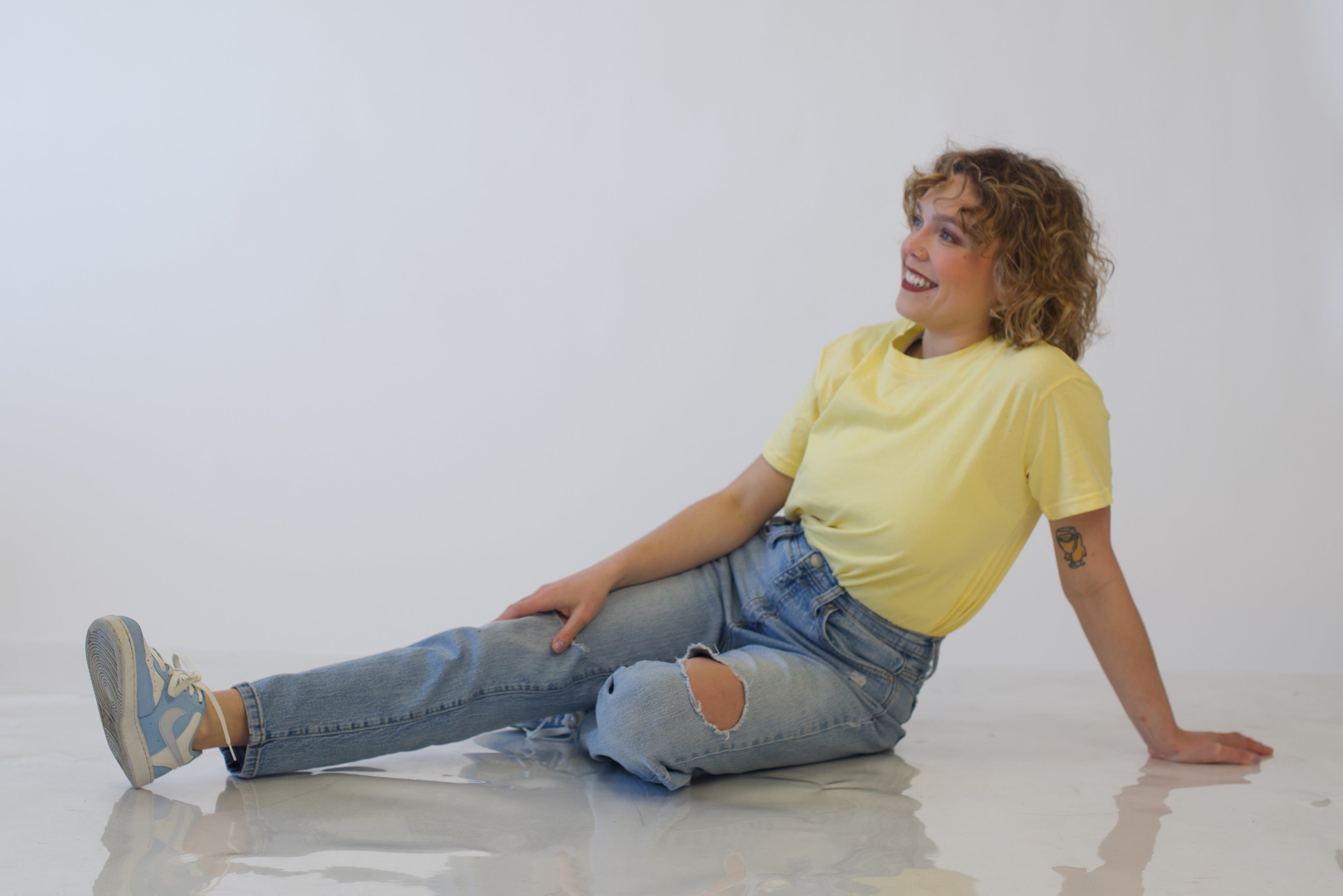 Person sitting on a white floor wearing a yellow shirt and blue jeans.