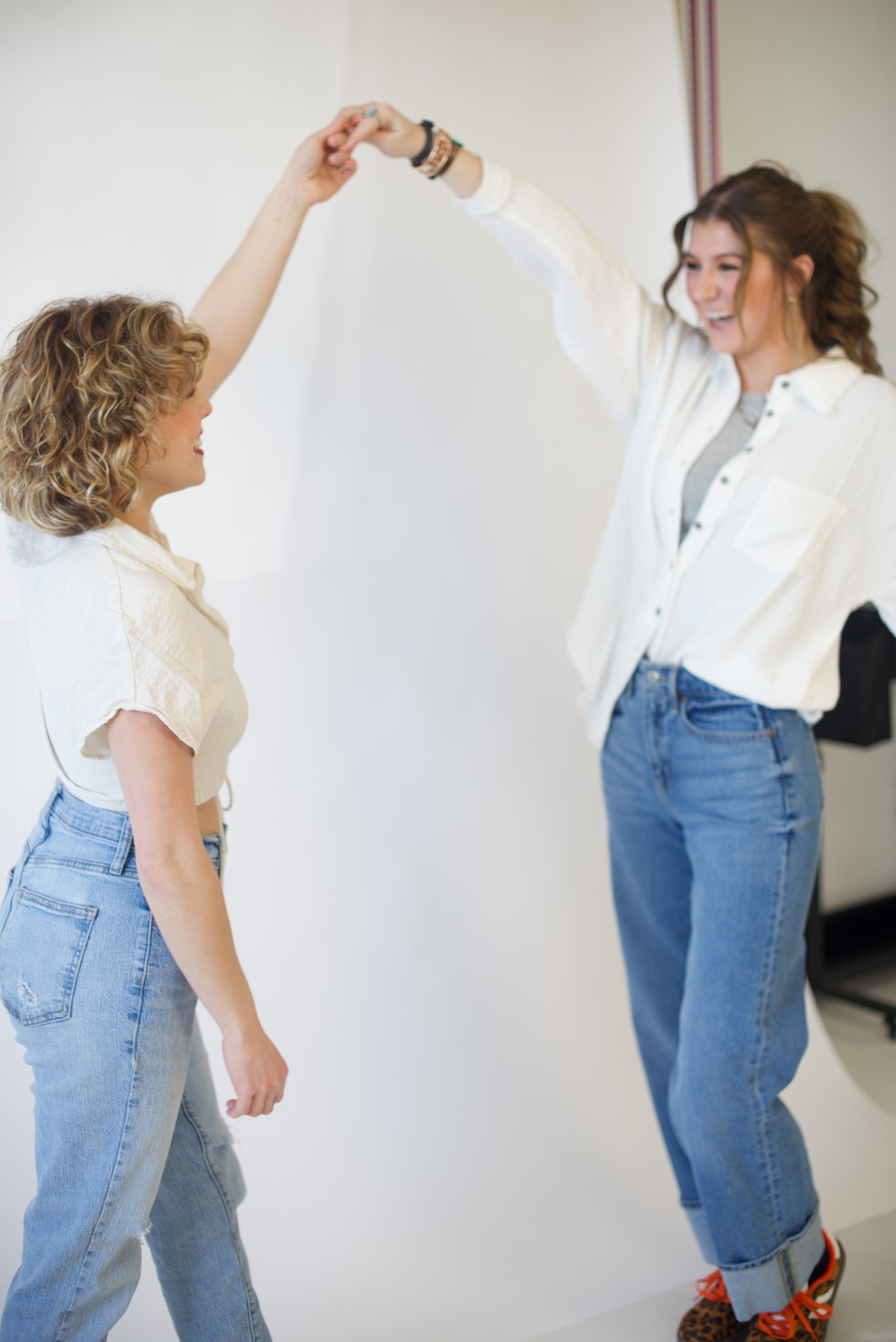 Two women dancing with each other, one spinning the other in white shirts and jeans against a plain background. 