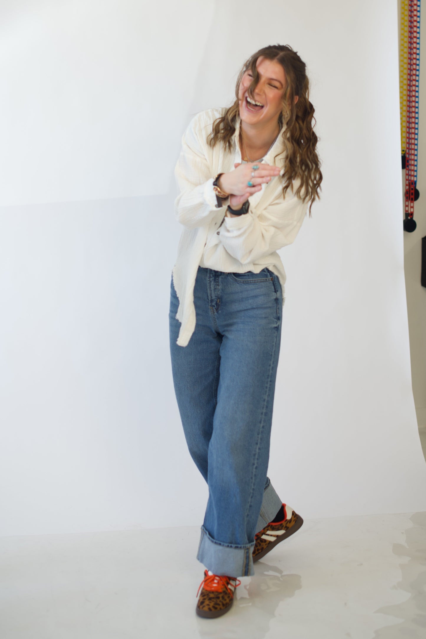 Person wearing a white sweater and blue jeans standing laughing in front of a white backdrop
